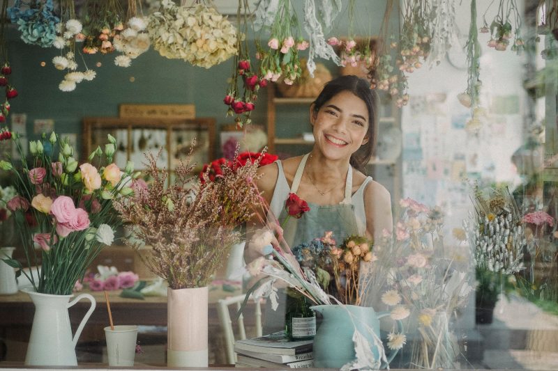 Through glass window of positive ethnic florist with dark hair standing near counter with flowers in floristry store in daytime