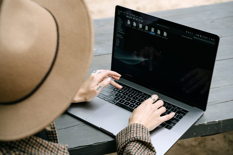 A person wearing a hat types on a laptop outdoors on a wooden table.