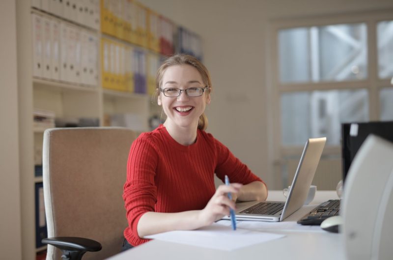 Australian Government Jobs for Teachers: Where Your Skills Actually Fit Happy woman in red sweater working at office desk with laptop and pen, smiling at camera.
